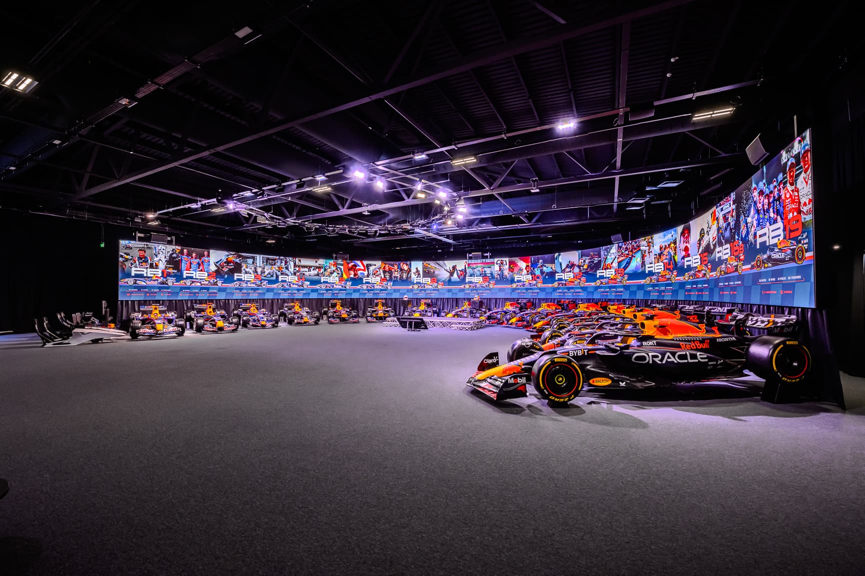 A brightly lit indoor display of Formula 1 cars lined up side by side, with colorful racing graphics on the walls above them.