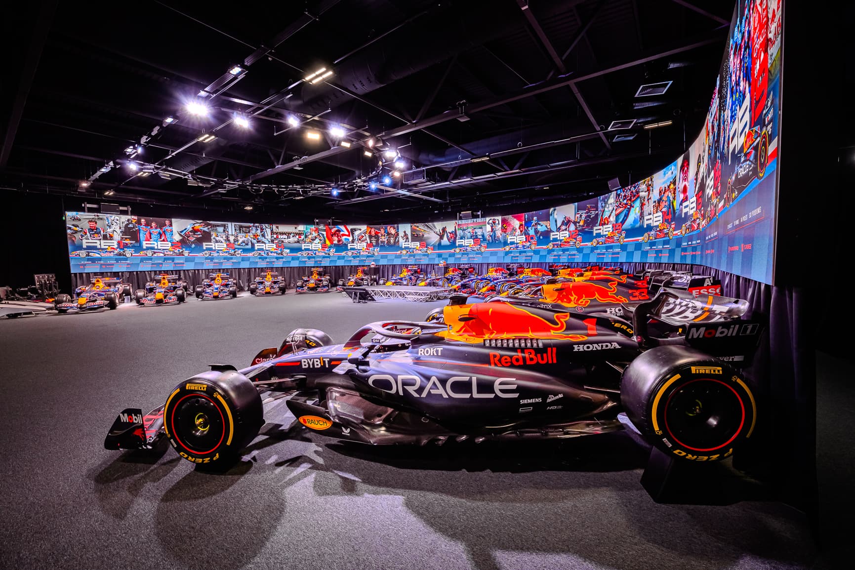 A display of Oracle Red Bull Racing Formula 1 cars in a brightly lit MK-7 event space with large screens showing racing highlights in the background.