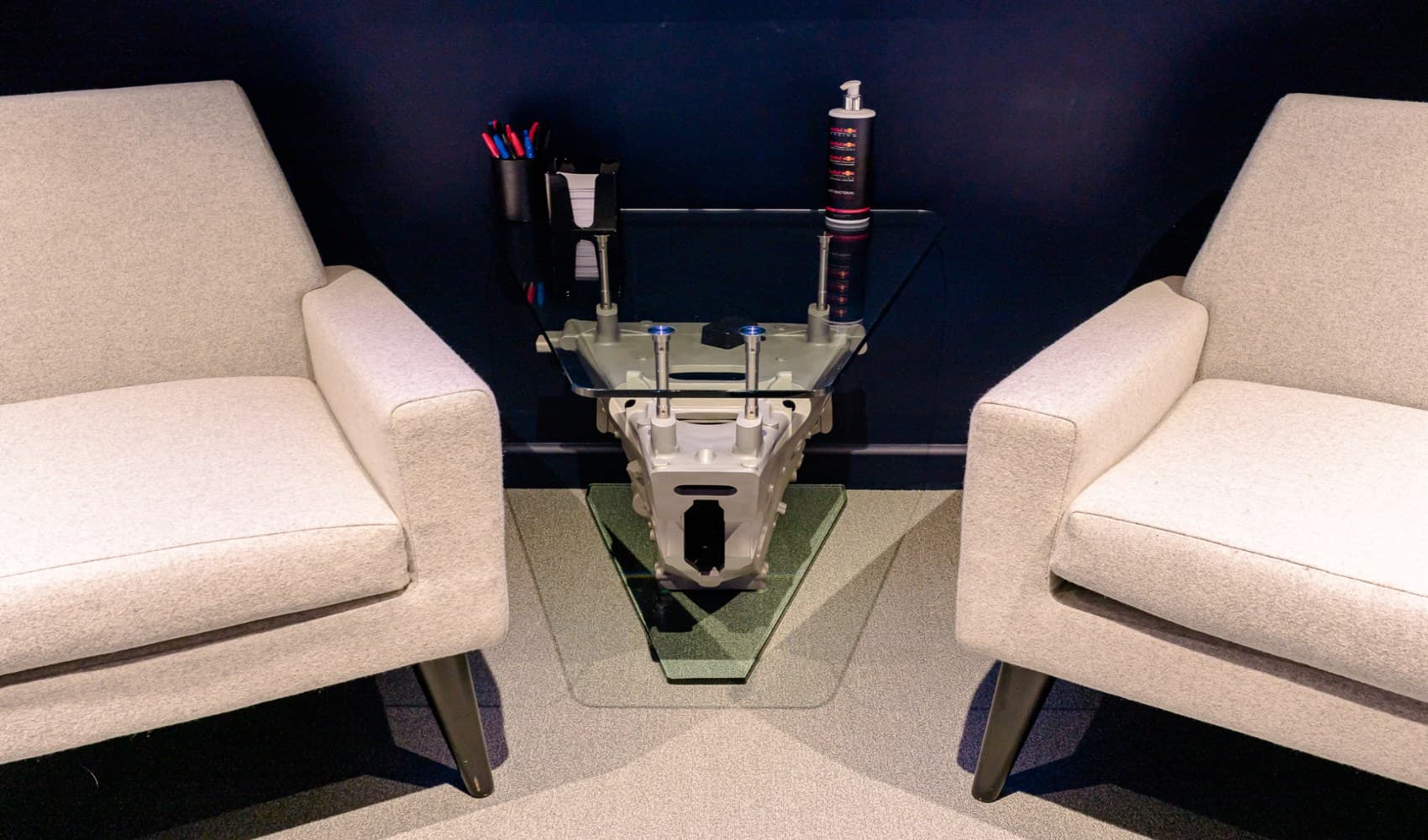 Two beige armchairs flank a unique glass coffee table featuring an engine block design, with pens and a racing damper on top.