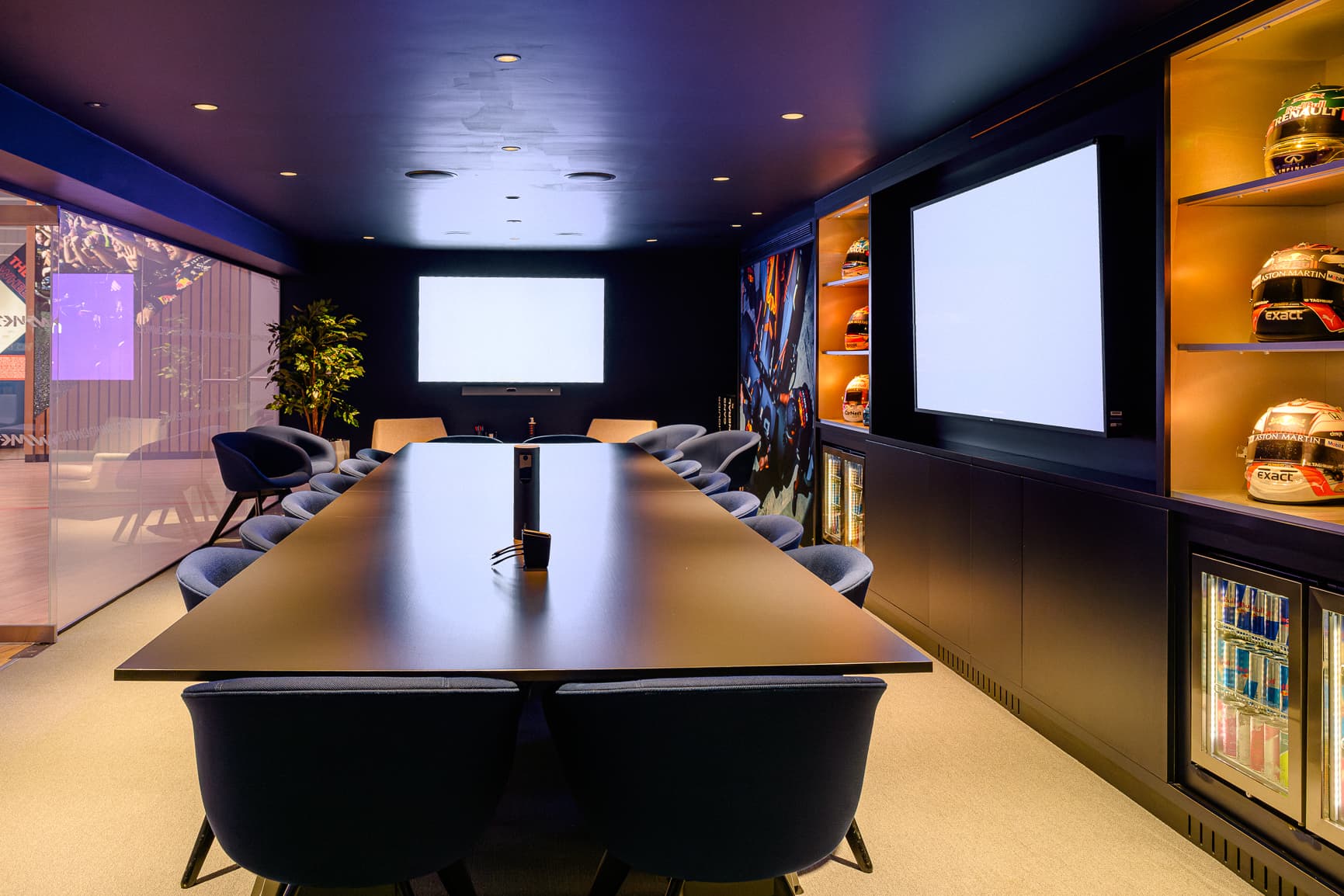 A modern conference room with a long table, blue chairs, racing helmets on shelves, and large screens on dark walls.