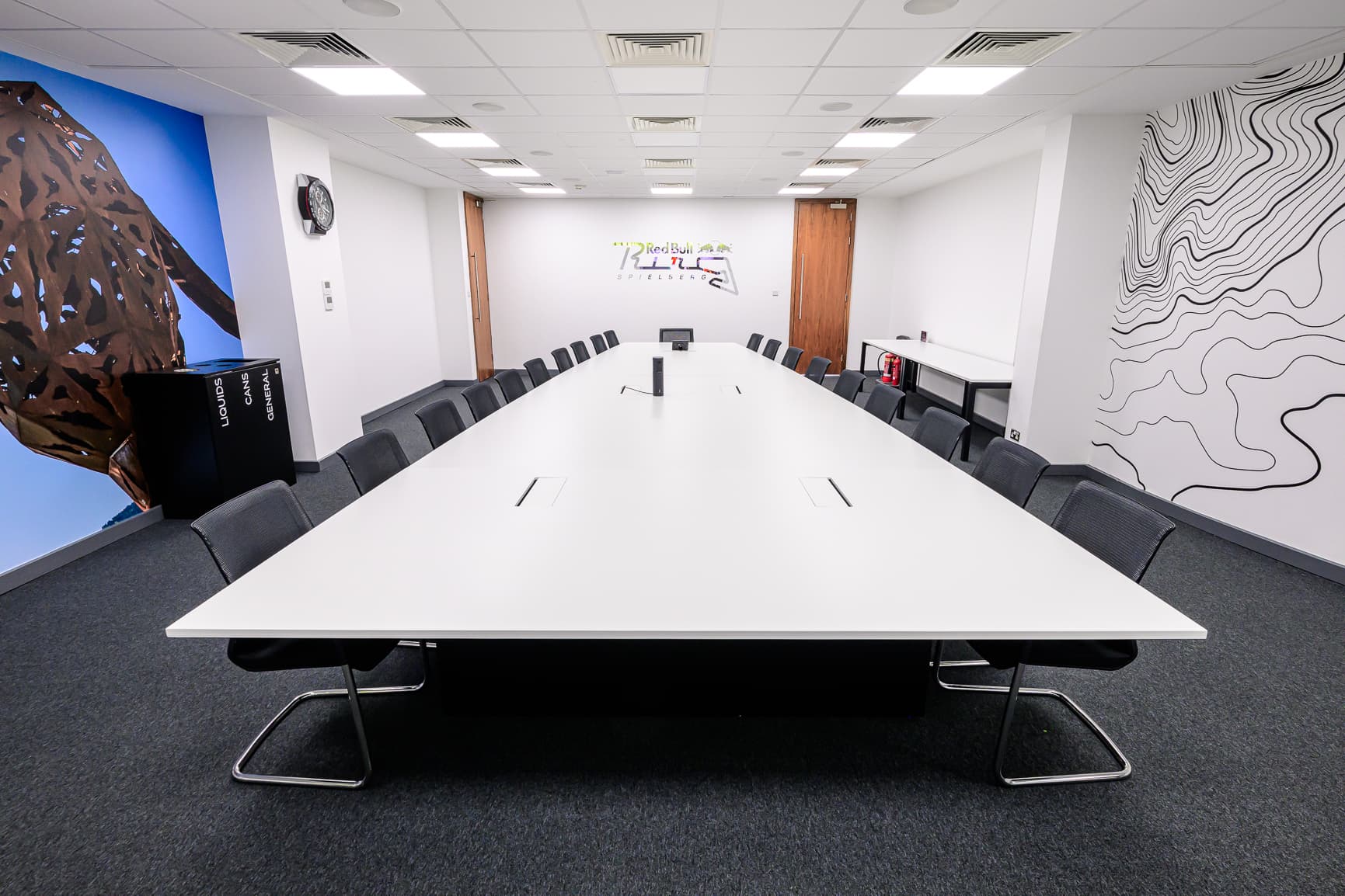 Modern conference room with a long white table, surrounded by chairs, abstract wall art, and a colorful logo on the back wall.