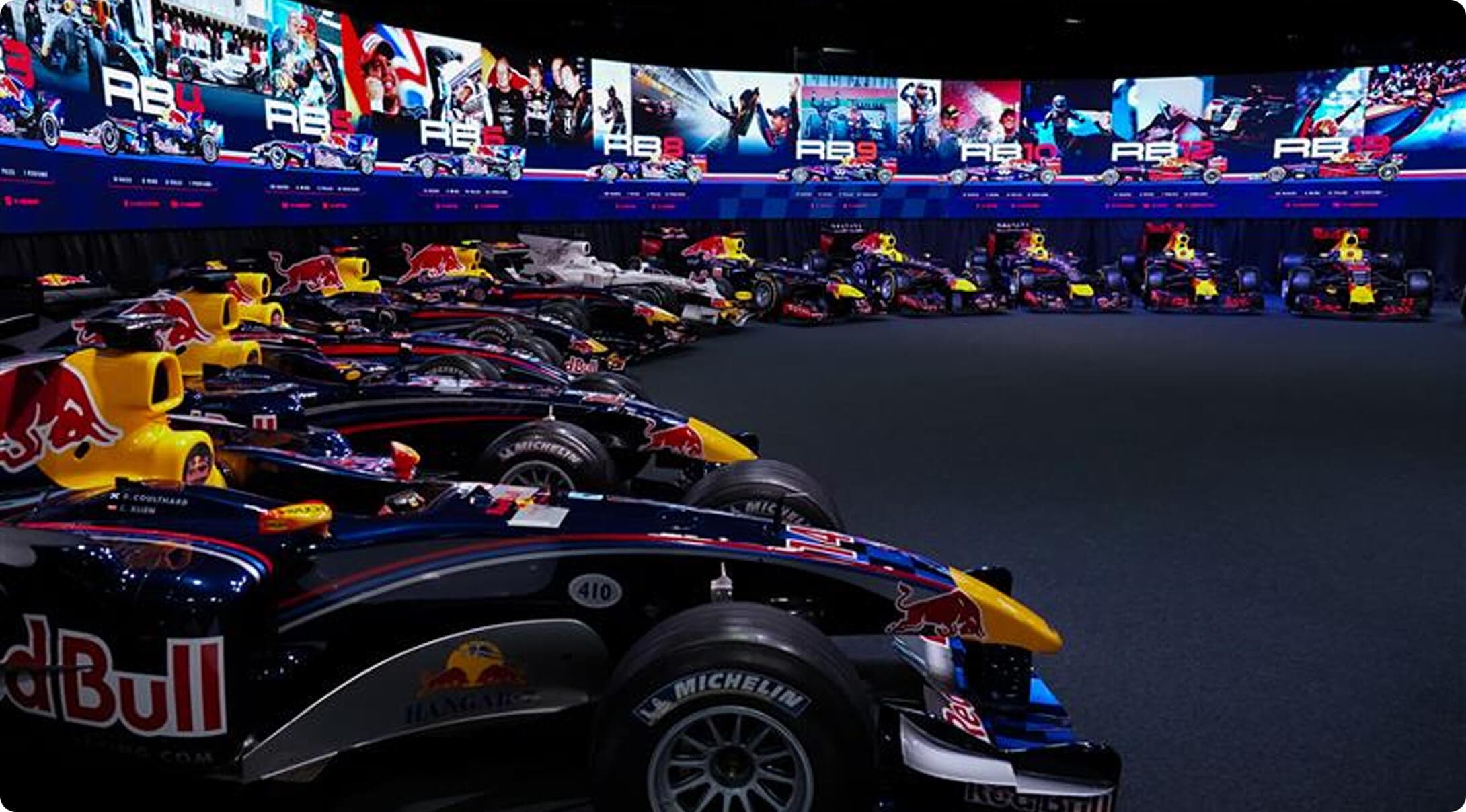 A row of Red Bull Formula 1 cars displayed in our MK-7 Event Space, with a large screen above showcasing images and info related to Red Bull's racing history.