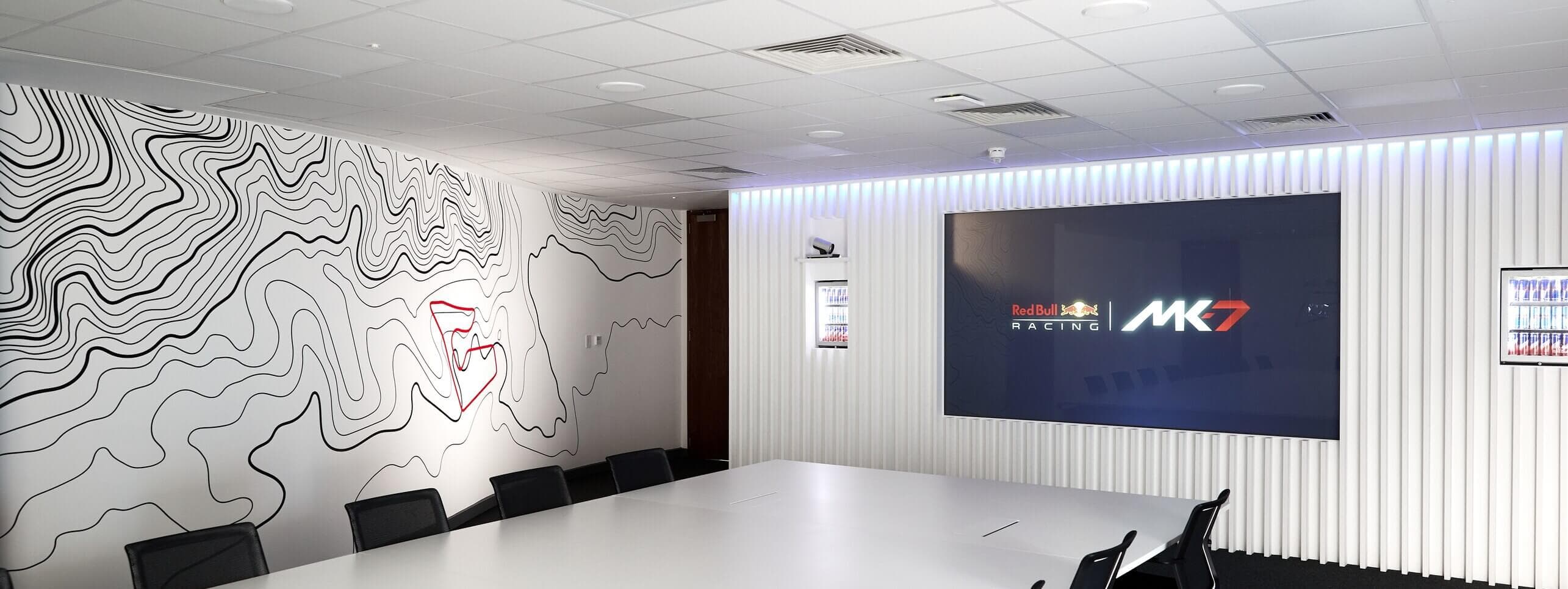 Modern conference room with a large display featuring "Red Bull Racing MK7," abstract wall art, white table, and black chairs.