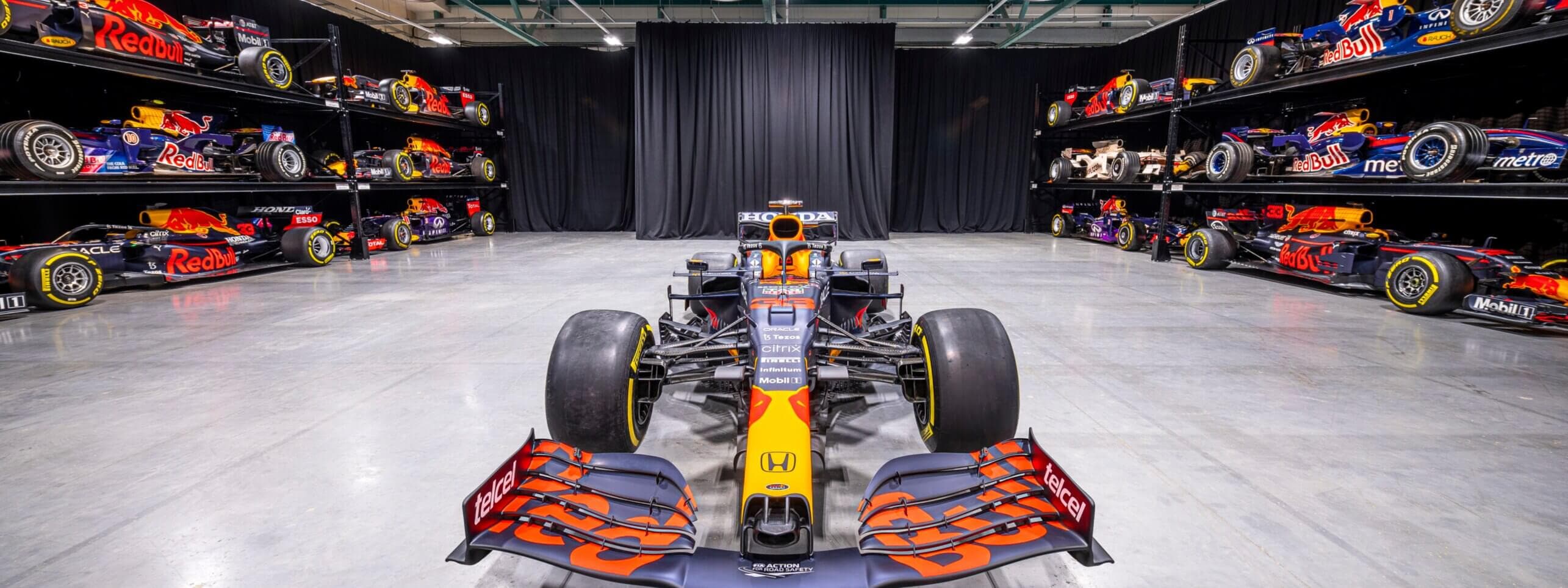 F1 car in a showroom surrounded by other Red Bull racing cars on display shelves, with a concrete floor and black curtains.